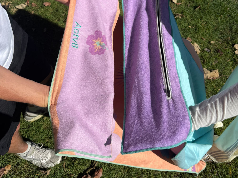 Towels held up on the grass, a lilac one with a purple flower and the Actv8 name and a purple one with a zip pocket