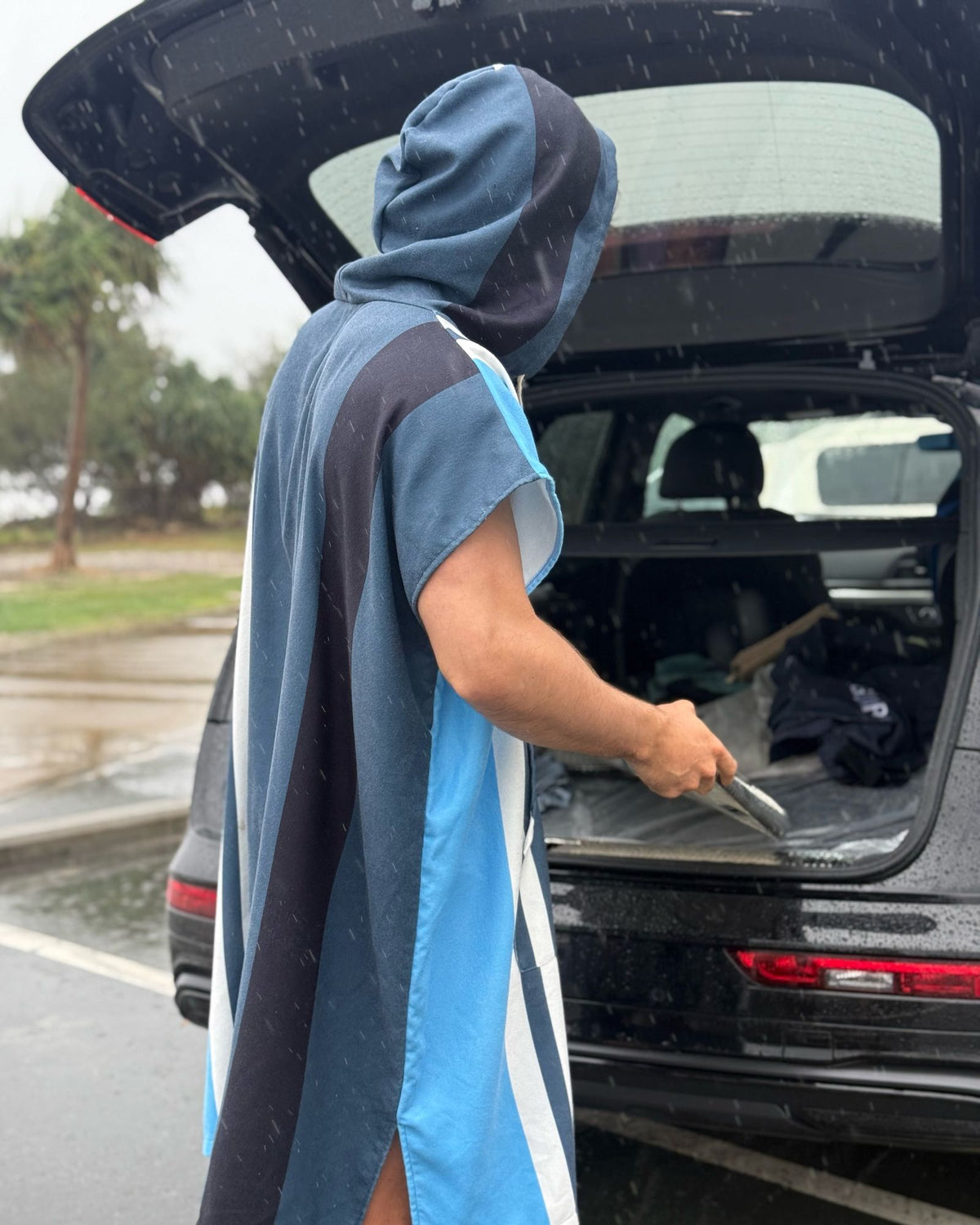 Changing Poncho in Coda, worn with the hood up at the open boot of a car in the rain