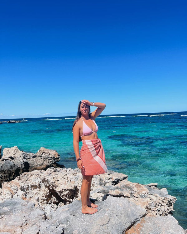 Woman on coastal rocks by turquoise water with a pink towel wrapped as a sarong