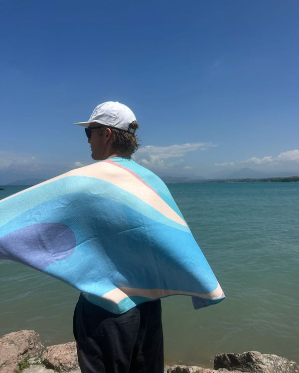Man in a white cap holding a blue towel around his shoulders beside a lake
