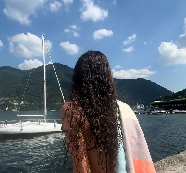 Woman with long curly hair and a towel over her shoulder looking out at a lake and sailboat