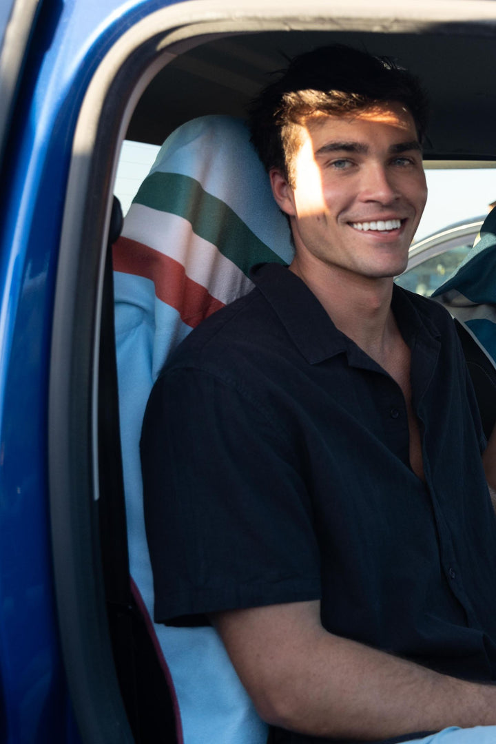 Smiling man sitting in a blue car, a striped Actv8 car seat towel covering the seat behind him