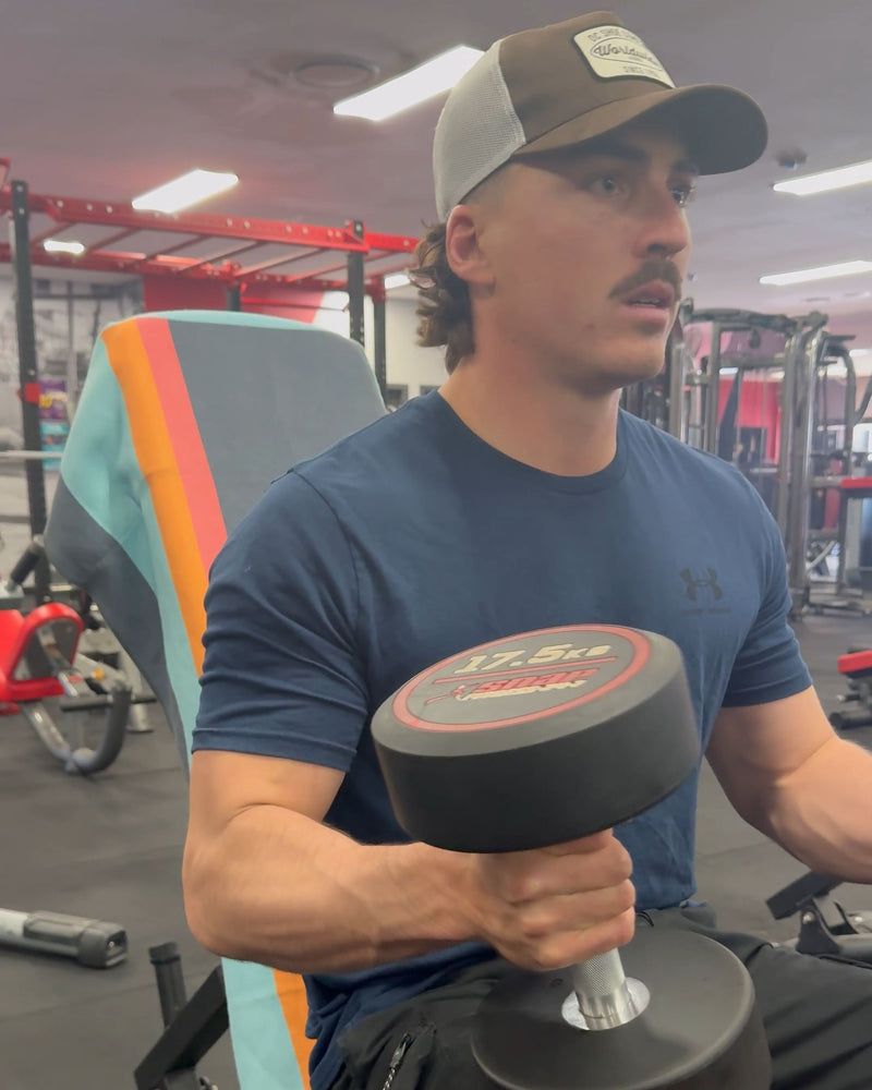 Man doing dumbbell curls in the gym, sitting on a bench with a striped Actv8 towel over the backrest